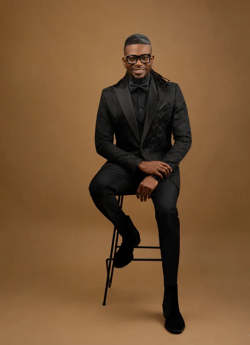 Toyin Adeniji, Ph.D., seated on a stool in a black jacquard tuxedo with a black paisley bow tie and pocket square, smiling, against a warm brown backdrop